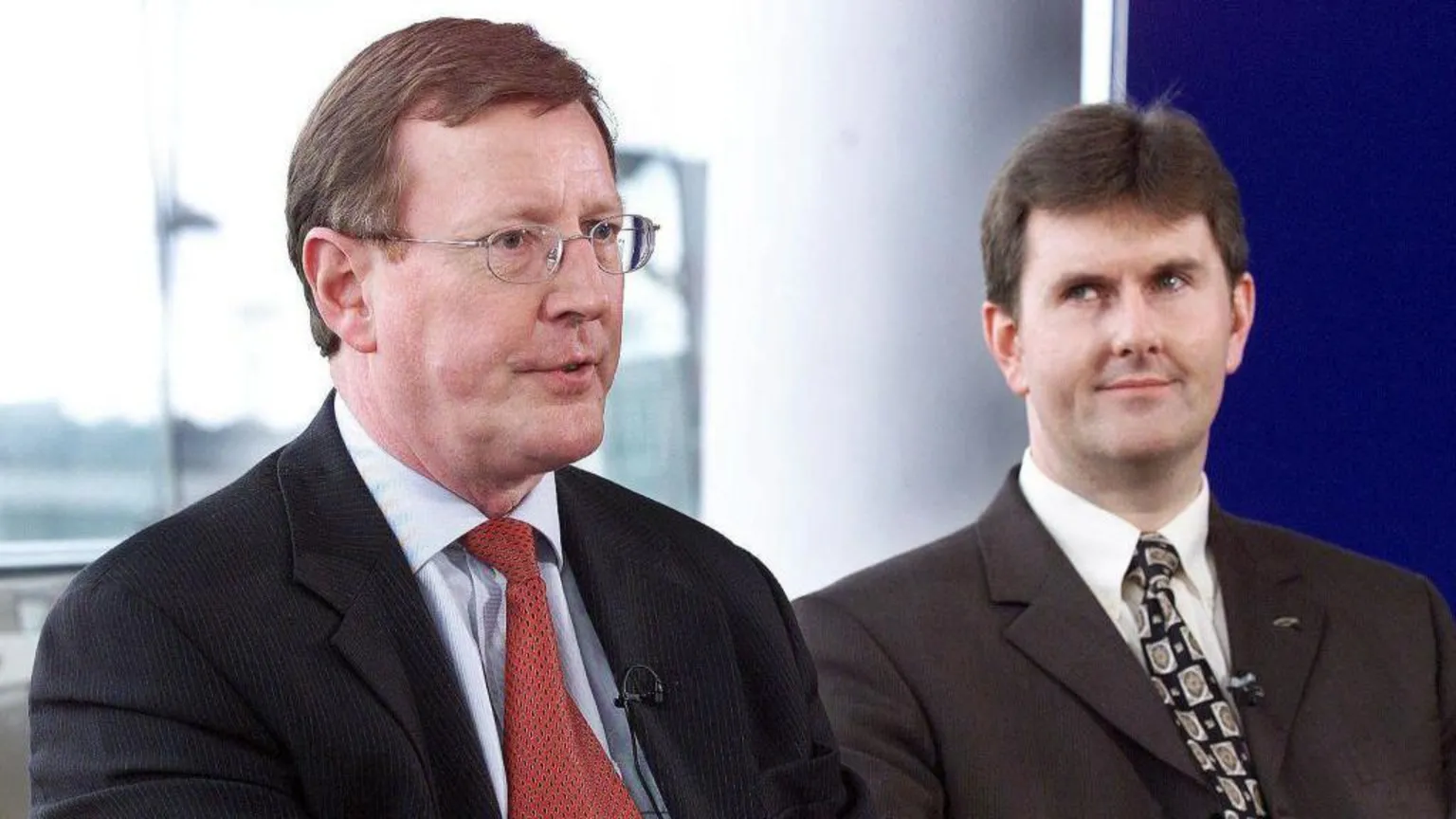  An older photograph of two men. The older man on the left has short brown hair and is wearing small metal framed glasses. He is wearing a dark suit, light blue shirt and red patterned tie. The younger man on his left is looking at the older man with a raised eyebrow and slight grin. He he has black hair and is wearing a dark suit, white shirt and black patterned tie.
