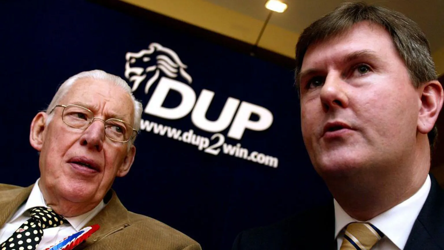  A close up image of two men. The older man on the left is looking at the younger man on the right. The older man has slicked back white hair and is wearing metal square framed glasses. He is wearing a light brown suit, white shirt and a black and white spotted tie. He is also wearing a red, white and blue rosette. The younger man has black hair and is wearing a dark suit, white shirt and a brown, white and black striped tie. There is a large DUP sign in between them.