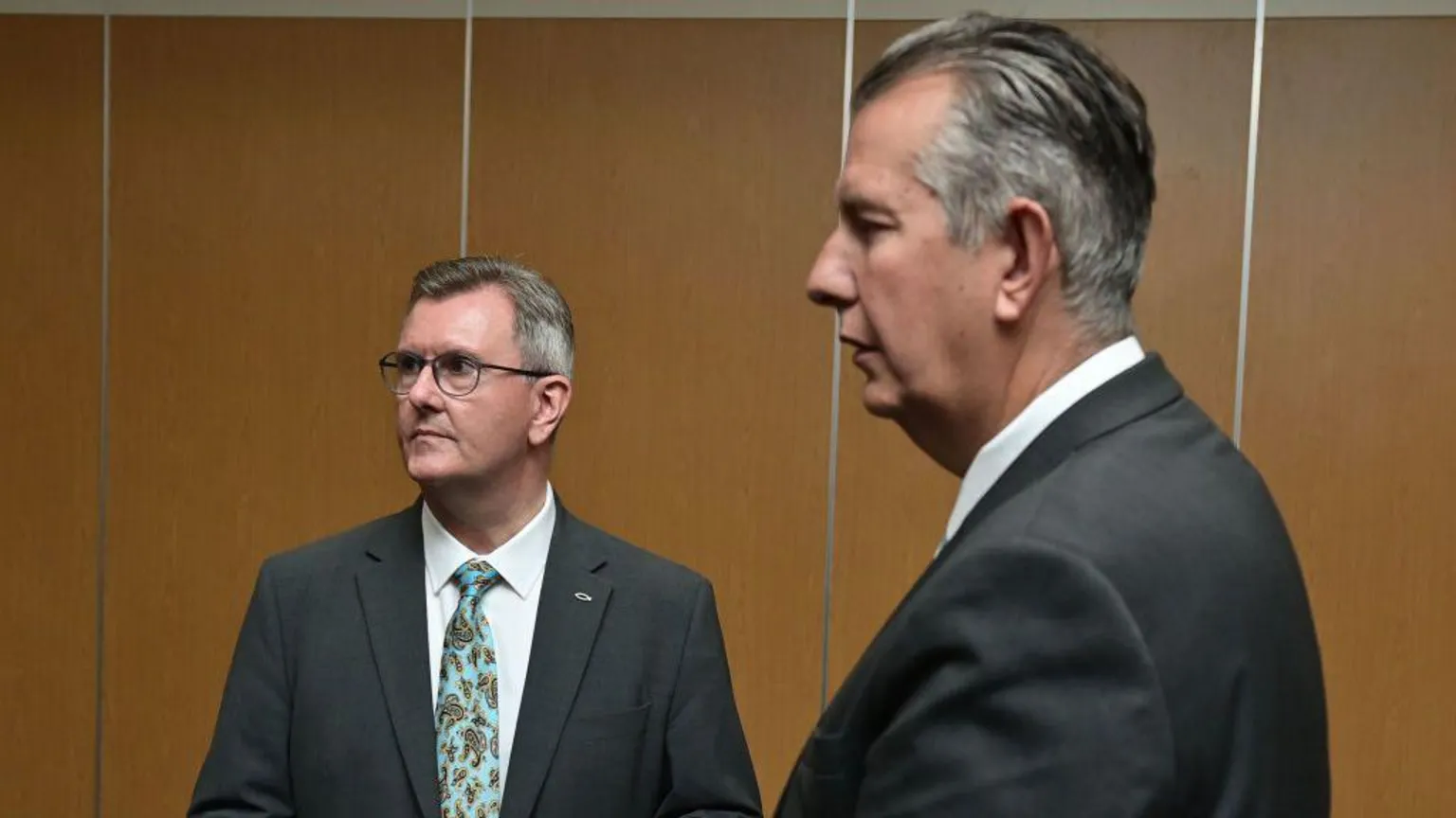 Two men look off to their right. In the foreground is a tall man with slicked back grey and white hair. He is wearing a dark suit and white shirt. The shorter man in the background can be seen more visibly. He is wearing a grey suit, white shirt and turquoise patterned tie. He is also wearing glasses and has a fish badge pinned to his jacket. They are both standing in front of a brown wall.