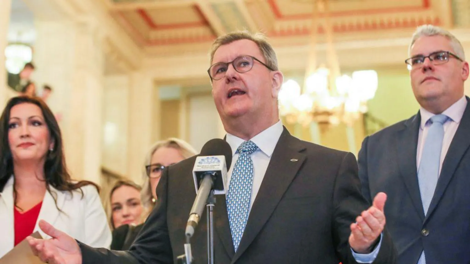  A man speaks into a microphone. He has grey hair and is wearing glasses. His arms and hands are stretched out. He is wearing a dark suit, white shirt and light blue patterned tie. He has a fish badge pinned to his jacket. The man on his left is taller and has white hair. He is wearing glasses, a grey suit, light blue tie and white shirt. The woman on the left of the image has long dark hair. She is wearing a white jacket and red top underneath. The background is blurred.
