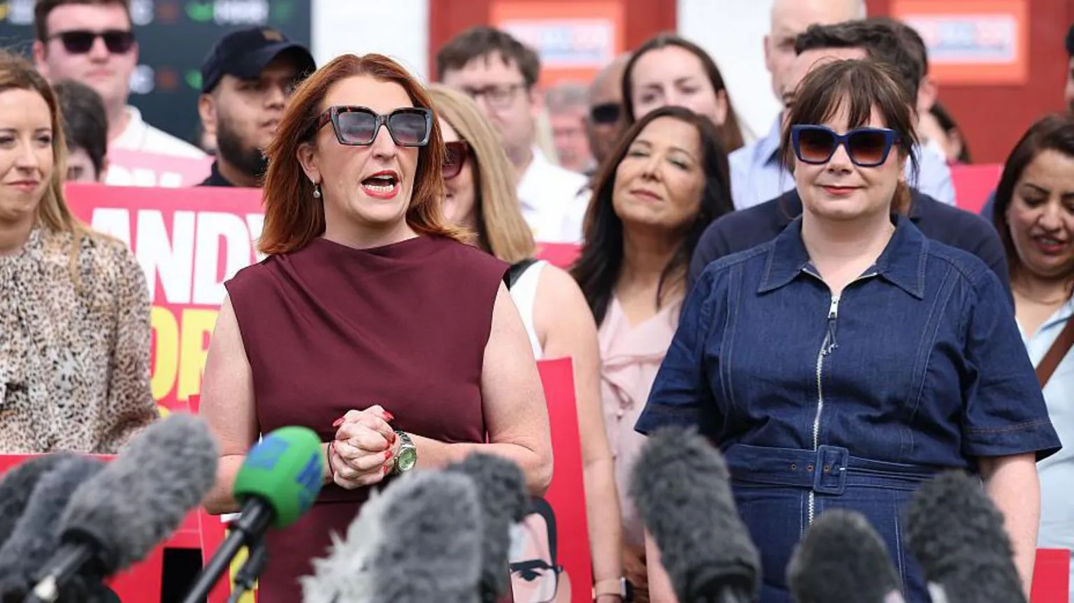 Getty imges Louise Haigh, a woman with shoulder length ginger hair and sunglasses, addresses the crowd at Andy Burnham's by-election victory rally, as Anneliese Midgley, in a blue denim dress and sunglasses, looks on