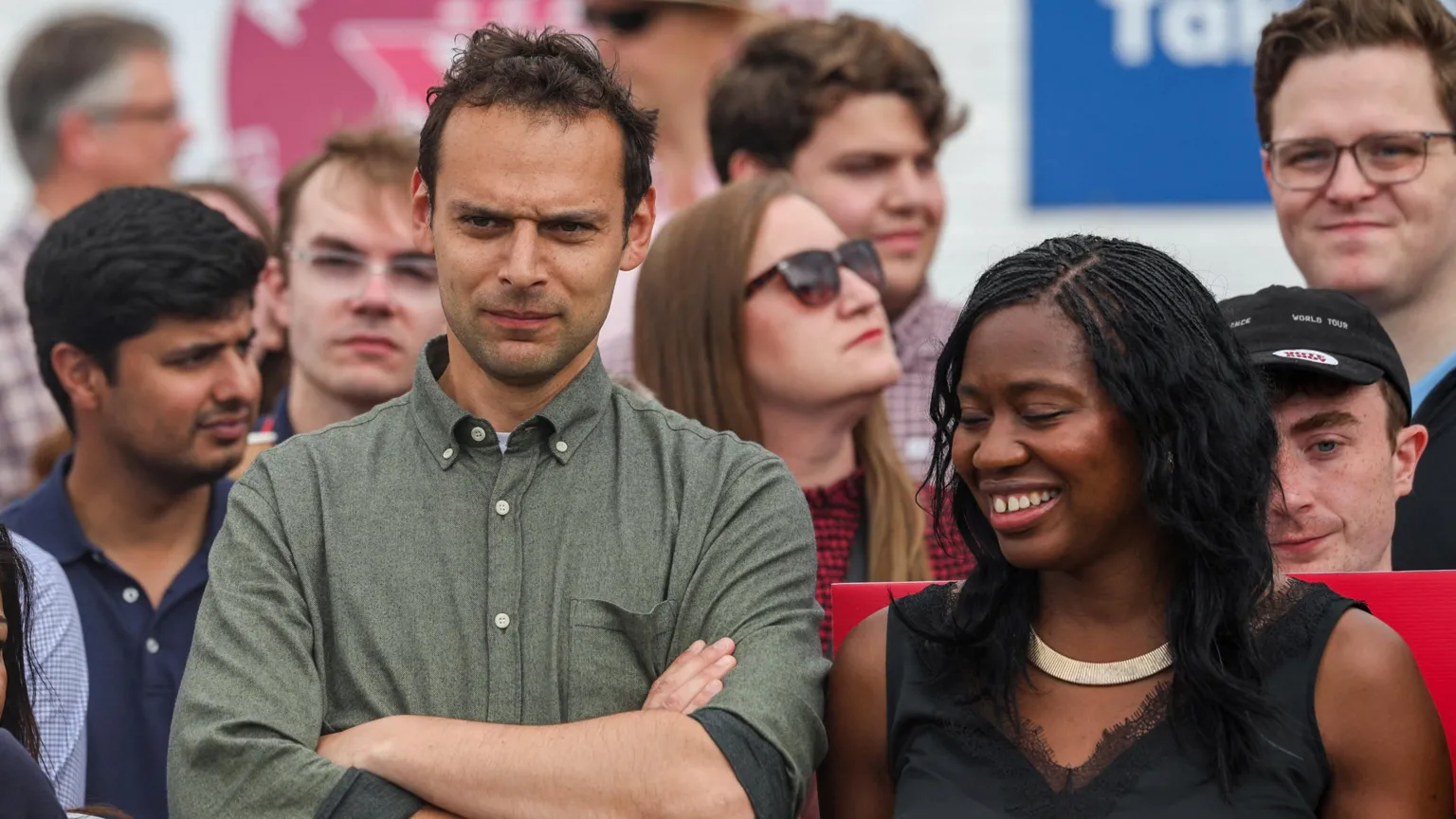 Josh Simons, in a green shirt with a button down collar, folds his arms and looks on as Burnham delivers a speech to supporters. A smiling Miatta Fahnbulleh, in black top, is stood next to him