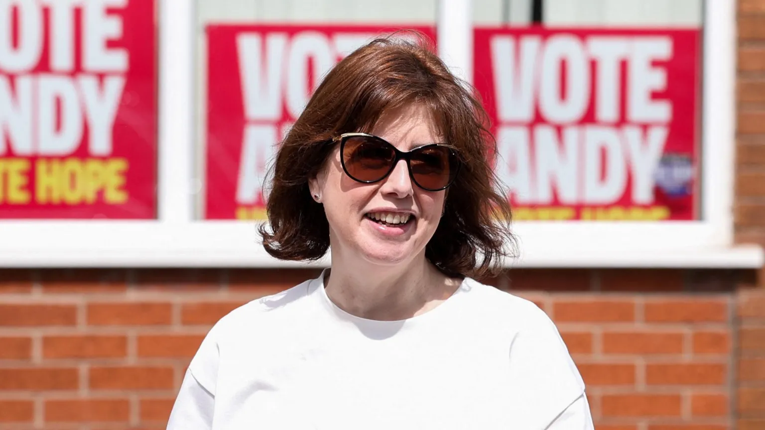 Lucy Powell, a woman in white T-shirt, with shoulder length hair and sunglasses, pictured against a backdrop of Vote Andy posters