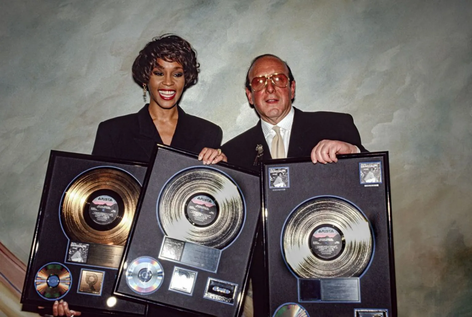 Whitney Houston and Clive Davis hold up a collection of gold and platinum records, celebrating the success of her album I'm Your Baby Tonight