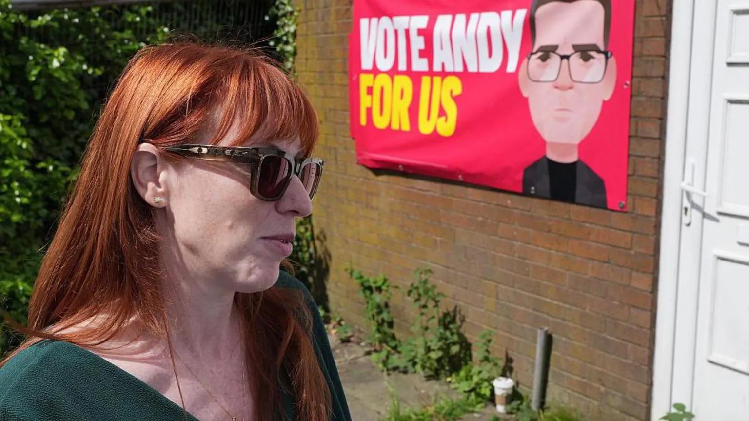 Angela Rayner, wearing dark glasses and a stern expression, against a backdrop of a large Vote Andy For Us banner