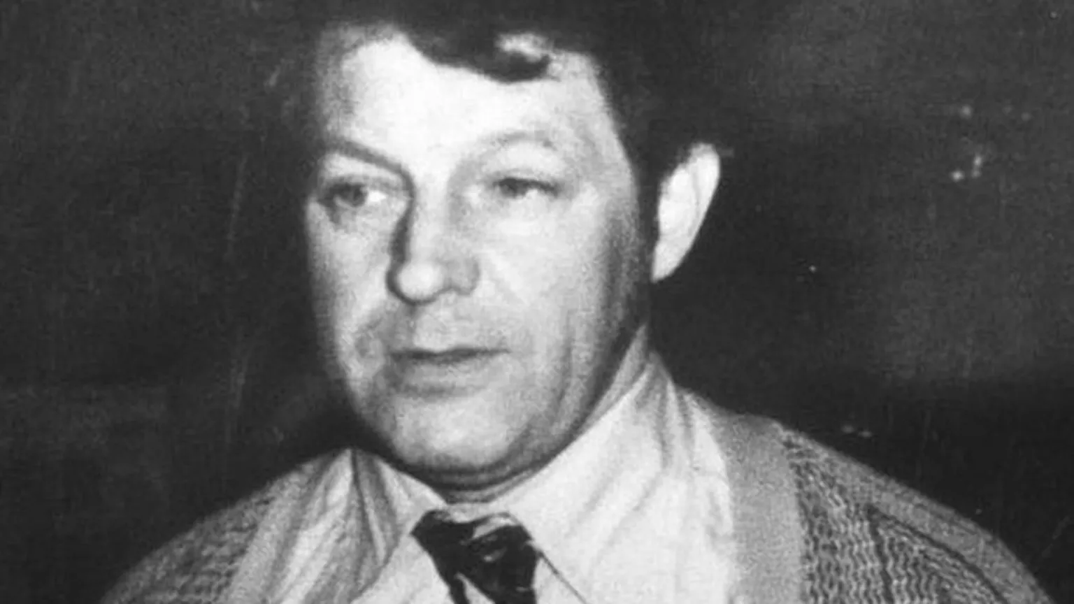 A black and white photo of a man with dark hair, wearing a shirt, tie and cardigan.