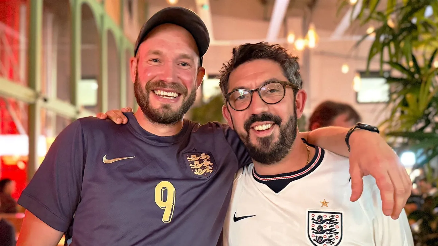Two fans, Tim and Dan, stand close together in the pub, with one person’s arm resting across the other’s shoulders as both face the camera. Each wears an England football shirt—one dark with a gold number “9” and crest, the other white with a crest and star above it. Both have facial hair and one wears a cap while the other wears glasses