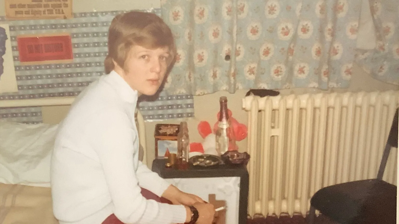 Pam Johnson A young woman with short hair sits on a bed, smoking a cigarette. The picture is taken in the 70s, so it looks old, and in the background you can see an empty bottle on a bedside table. The pattern on the curtains and the posters on th wall give the impression it is a teenager's bedroom.