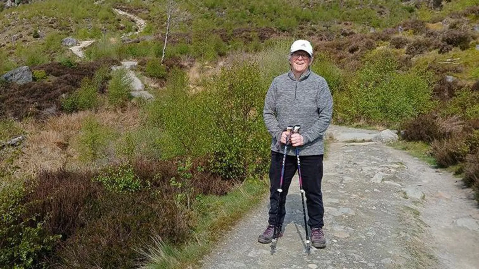 Pam Johnson A full-length photo of a woman posing on a rocky hill, the path in the background suggests she is high-up. She's wearing a grey fleece, black hiking trousers and walking boots, she's carrying tow poles to help her walk and a cap to keep the sun off her face. She's surrounded by shrubs and rocks.