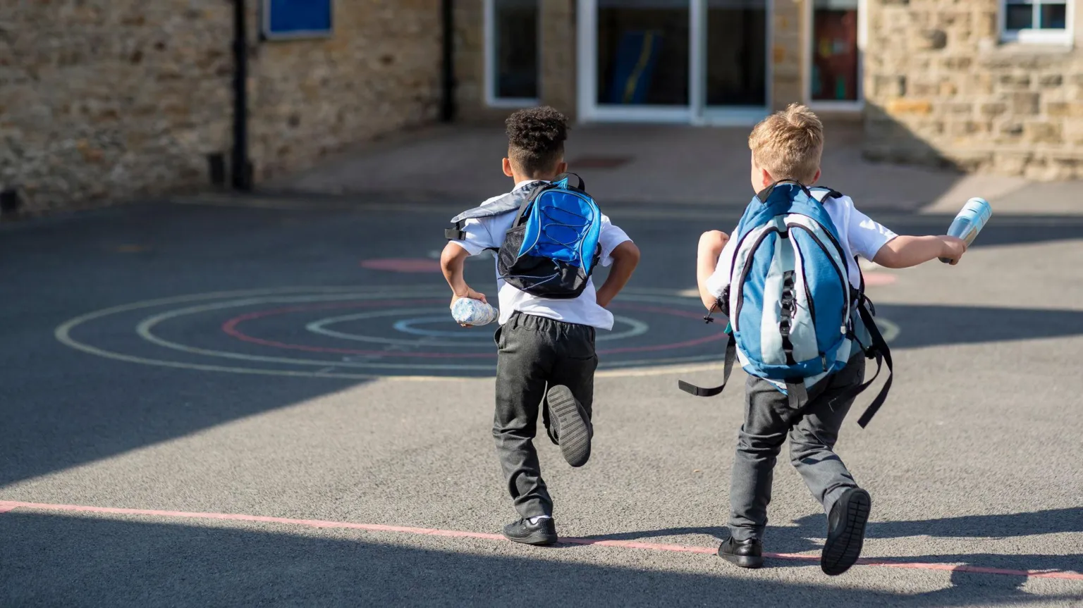/SolStock Two boys running in their school yard. They are running towards the door with their backpacks on.