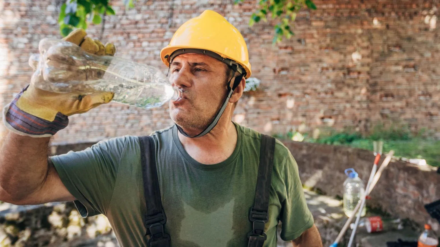 Man wearing a yellow safety helmet and sweaty green T-shirt drinking water from a plastic bottle