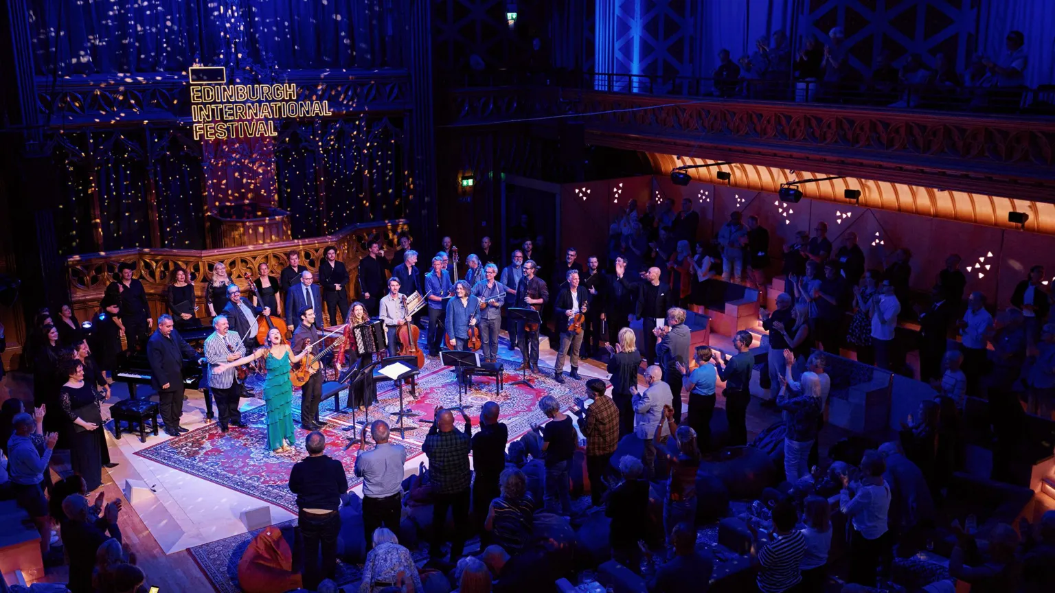 EIF Wide shot of a live performance at the Edinburgh International Festival inside a grand, historic hall, with a large group of musicians and singers on a central stage surrounded by a standing audience, ornate balconies and patterned walls lit in blue and warm tones, and a glowing sign reading “Edinburgh International Festival” above the stage.