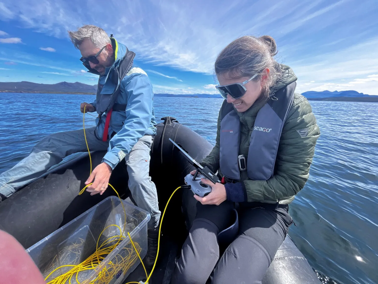 A woman with dark hair and sunglasses holding a controller with a yellow cable coming out of it into a box. A man is then feeding the cable into the sea. They are on a small boat on a clear day with blue skies.