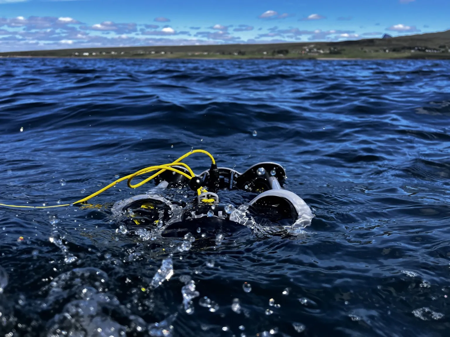 A small underwater drone sits on the surface of the sea, attached by a yellow cable. The land is seen in the distance.