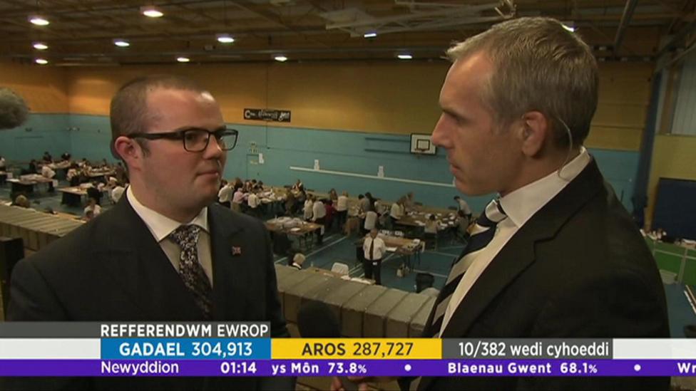 Mae'r llun yn dangos Llŷr Powell yn cael ei holi gan ohebydd. Mae'r ddau'n sefyll uwchlaw neuadd chwaraeon ble mae pleidlieisiau yn cael eu cyfrif.