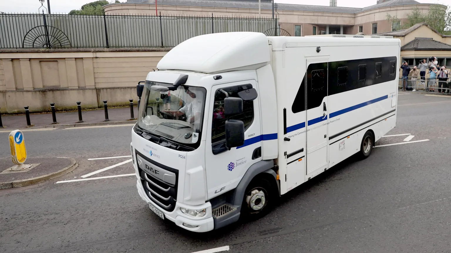A white prison service van driving away from court. Photographers can be seen the background taking pictures.