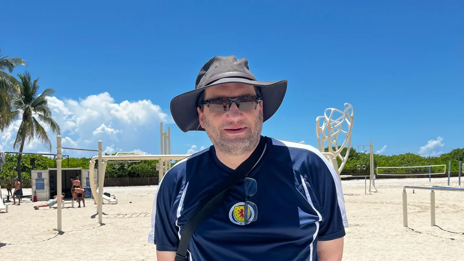 A man wearing a black hat and sunglasses and a Scotland football top. He is standing in front of a beach.