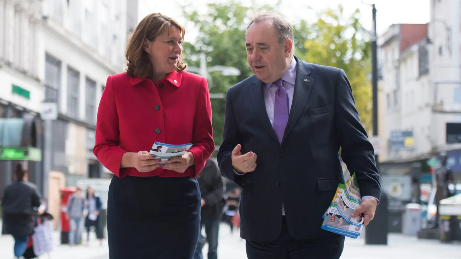 Two people in the centre of the photograph are talking and holding leaflets. To the left is Leanne Wood, who is wearing a dark skirt and a red jacket, and to the right is Alex Salmond, who is wearing a suit, a pink short and purple tie.