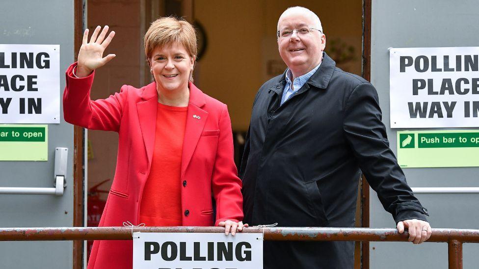 Nicola Sturgeon, who has short fair hair, and Peter Murrell, who is bald, standing smiling outside a polling station while leaning on a hand rail. Sturgeon, on the left, is wearing a pink jacket and red top. Murrell is wearing a dark overcoat.