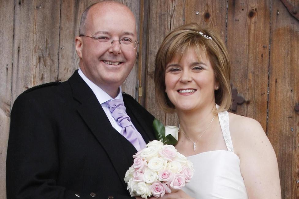 Murrell and Sturgeon in wedding attire posing in front of a wooden backdrop. He is wearing a dark suit with a light tie and buttonhole and she is in a white dress holding a bouquet of pale pink and white roses.