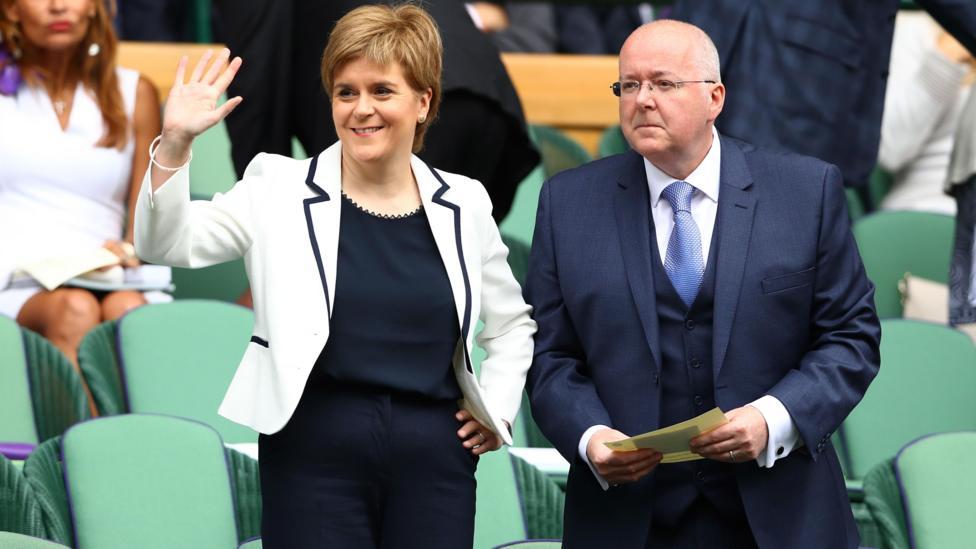 Sturgeon and Murrell at Wimbledon in 2016. They are standing among rows of green seats; She is wearing a white jacket over a dark outfit and raises a hand in greeting, while he wears a dark suit and holds a paper programme.