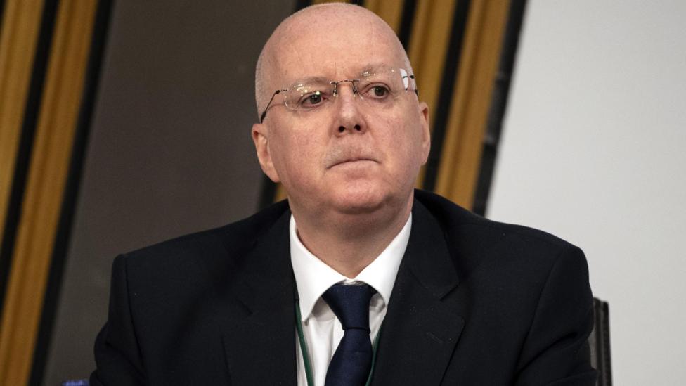 Close-up of Peter Murrell wearing glasses, a dark suit, white shirt, and tie, seated indoors in a Scottish Parliament committee room against a backdrop of curtains, looking slightly upward.