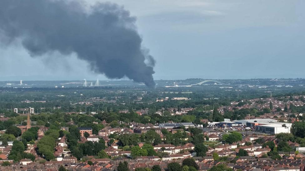 A large plume of smoke can be seen billowing up into the sky, with the Runcorn bridge visible in the distance.