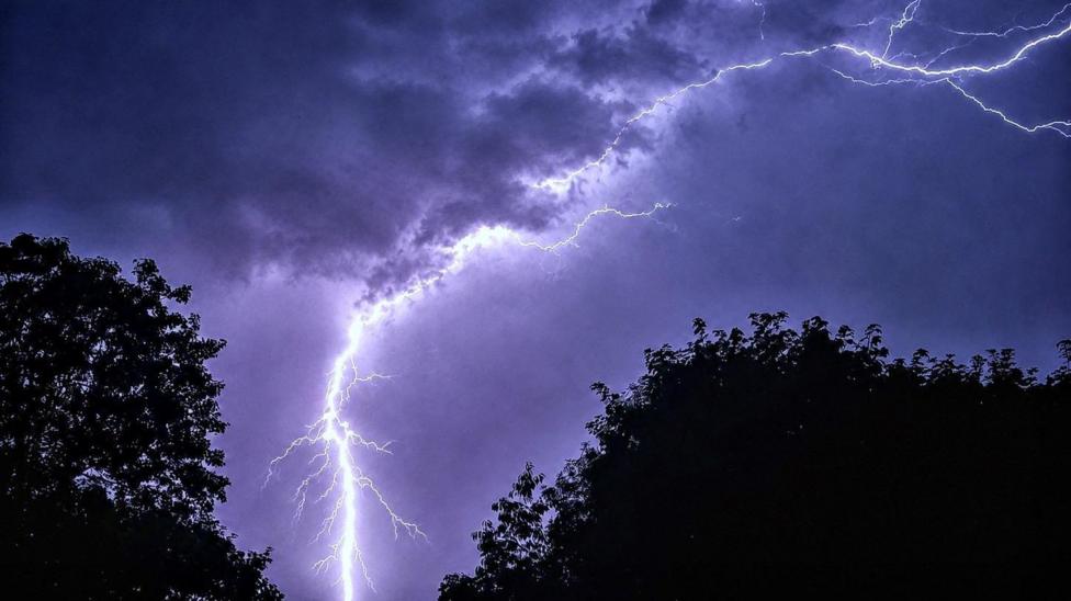 Lightning forks across a dark, stormy sky above silhouetted trees, illuminating thick clouds during a night-time thunderstorm.