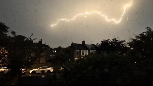 Lightning stretches across the sky above houses and a parked car during a heavy night storm.