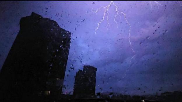 A flash of lightning lights up the sky behind tower blocks, with raindrops visible on the window.