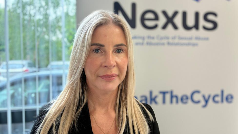 Joanne Barnes has long blond hair. She's dressed in black. A sign behind her states Nexus.