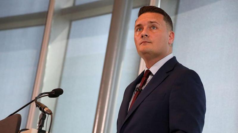 Wes Streeting stood at a lectern delivering a speech. He is wearing a navy blue suit, pale blue shirt, and red tie.