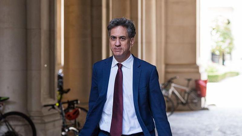 Ed Miliband walking down a cobbled pedestrianised street in London. He is wearing a white shirt, blue suit, and red tie.