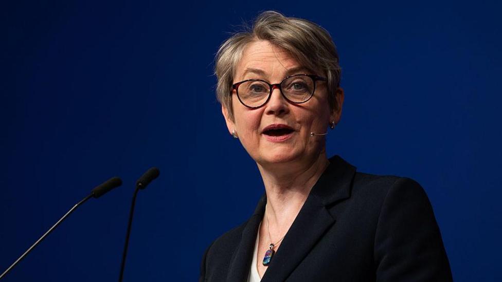Yvette Cooper stood in front of a lectern making a speech in front of a plain navy blue background. She is wearing a black jacket, necklace, and a white top.