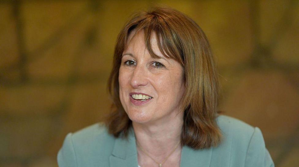 A smiling Rachel Reeves wearing a sky blue jacket and gold necklace with a top of a white top just visible in the bottom of the frame. Behind her is a mottled yellowish brown background.