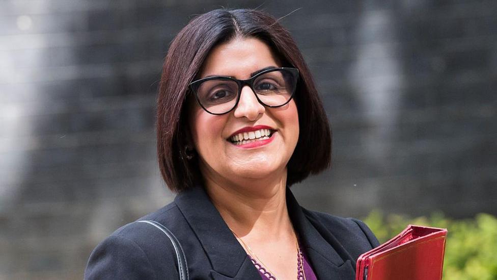 A smiling Shabana Mahmood stood in front of a black brick wall and a green bush in front. She is wearing a black jacket, purple top, and is holding a red folder.