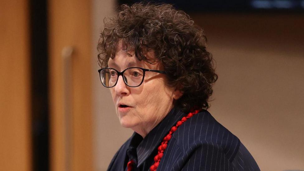 Elin Jones has glasses, brown curly hair, and wearing a blue pinstriped jacket.