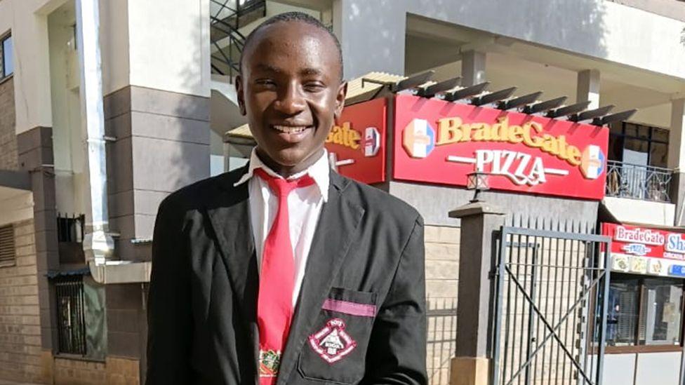 A photograph of Sylvester Muigai Ndung'u in a school uniform. He is in a grey jacket, white shirt and red tie.