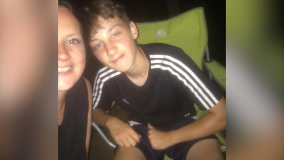 The photo shows Joshua in a black Addidas t shirt with white stripes on the shoulders. He is sitting in a green camping chair with a small smile on his face, posing for the photo next to his mum.