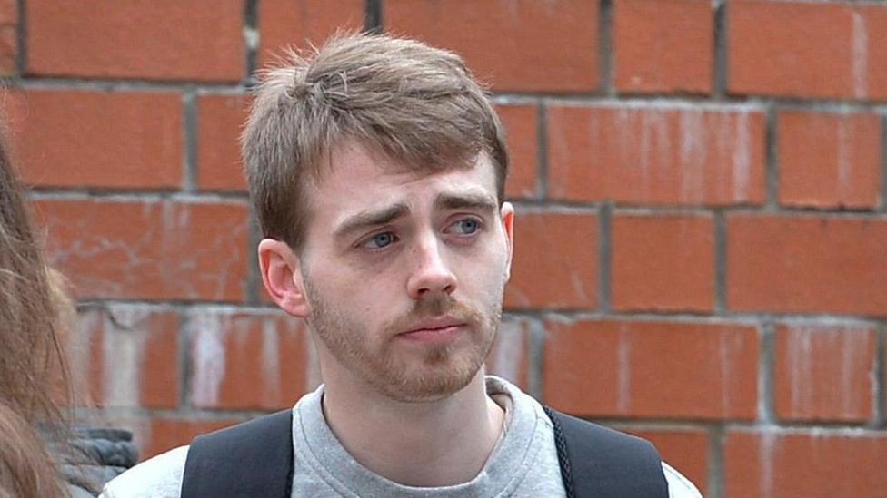 Kyle Waddell has short, brown hair and is wearing a grey sweatshirt with a rucksack over his shoulders. There is a red brick wall behind him.