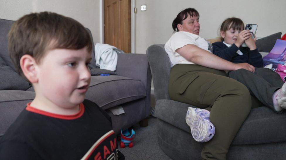 A picture of Carla, who has brown hair and is wearing a white top and green trousers, sitting in a chair with her daughter Jasmine. Her son is on the left.