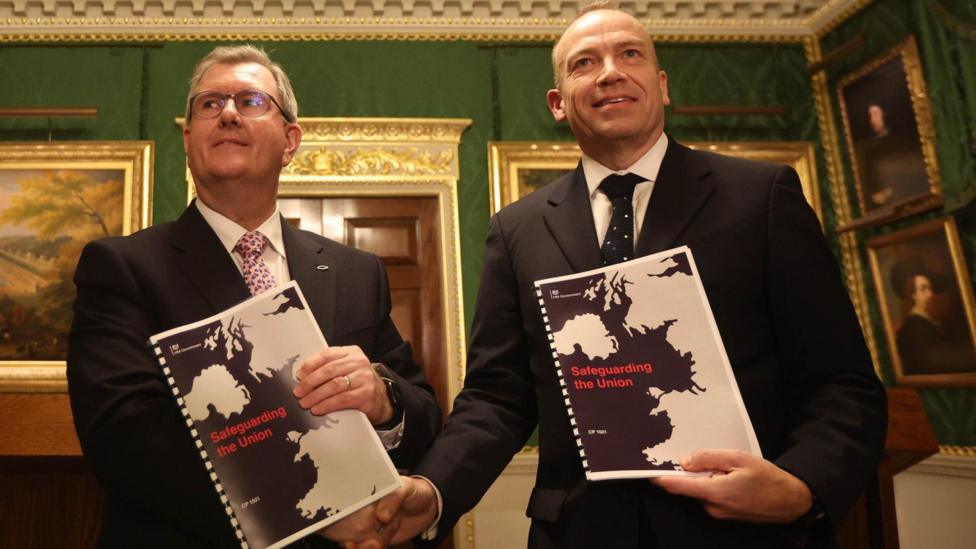 Jeffrey Donaldson and Chris Heaton-Harris shake hands and hold up a ring-bound paper entitled Safeguarding the Union. Both are in suits and are in a very formal-looking room with paintings in gold frames.