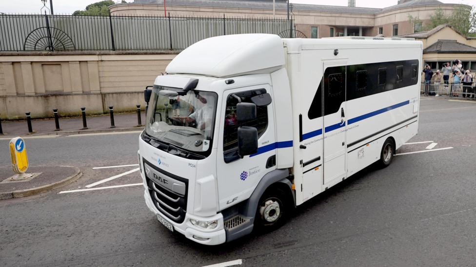 A white prison service van driving away from court. Photographers can be seen the background taking pictures.