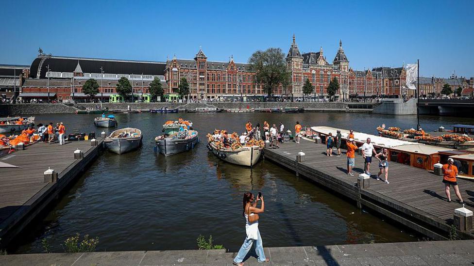 A woman walks past a canal in the Netherlands, where crowds of people are getting onto boats