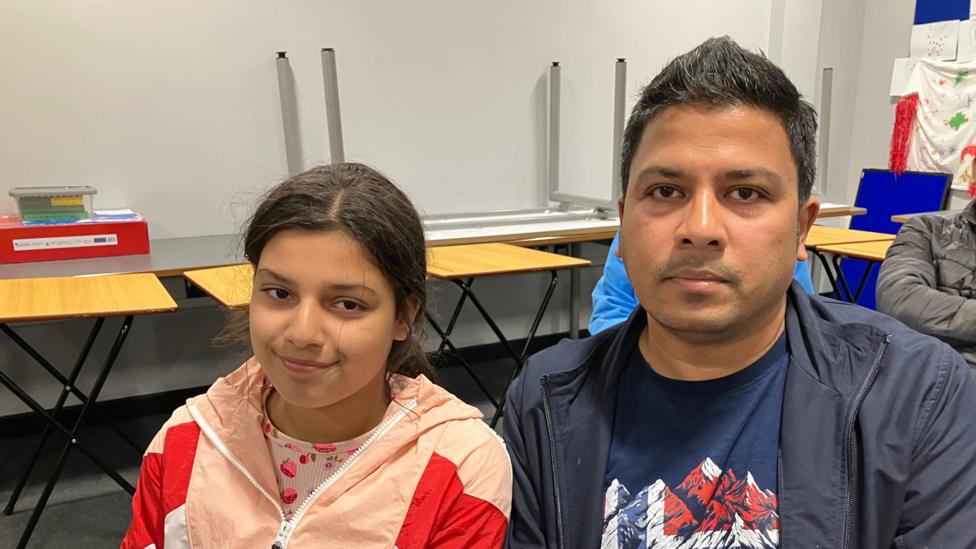 Ten-year-old Keisha is smiling at the camera and sitting next to her Dad, Abhishek, who is looking at the camera with a neutral expression. Keisha is wearing a top patterned with strawberries and a pink and red coat. Her dad is wearing a navy T-shirt and jacket. There are school tables stacked behind them.
