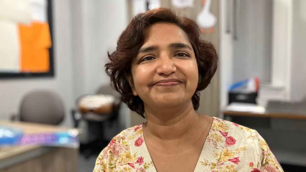 Aparna Nigam is smiling and looking at the camera. She is wearing a floral shirt. There is an out of focus classroom in the background.