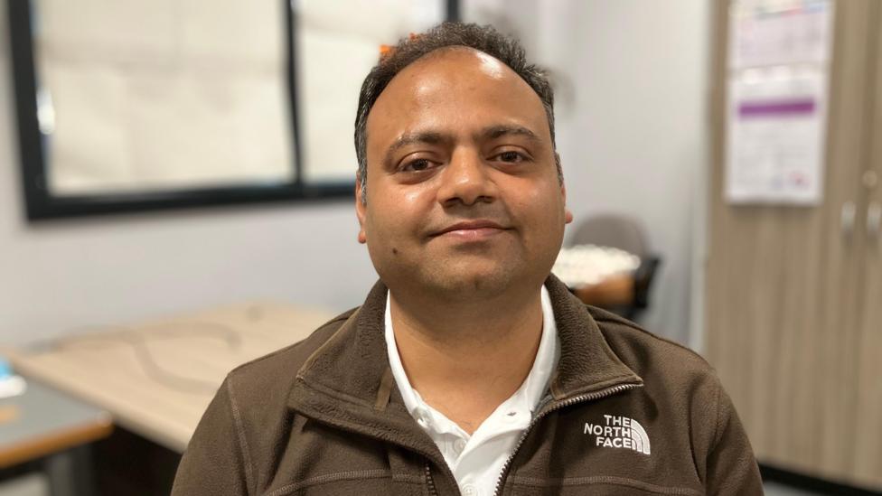 Abhishek Srivasdava smiling and looking at the camera. He is wearing a white shirt and brown fleece. There is an out of focus classroom behind him.