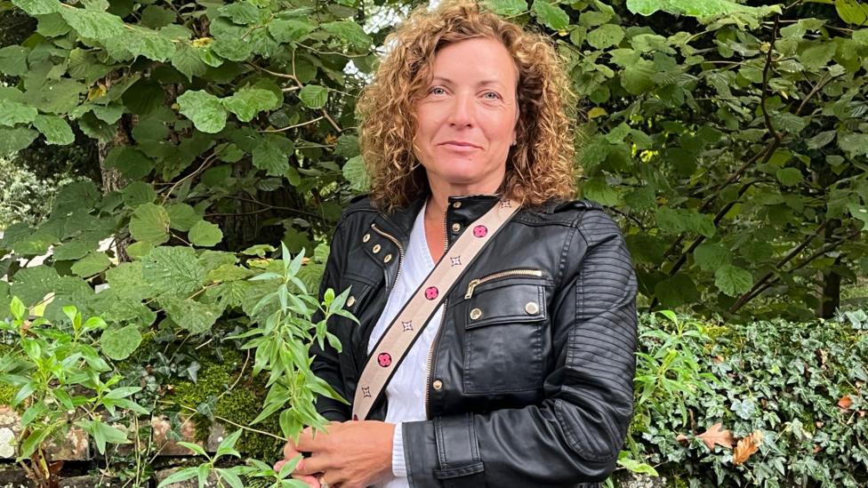 Sally Pavey has shoulder length curly hair and is standing outside surrounded by greenery. She is looking at the camera and wearing a black jacket and white top with a bag strap over her shoulder.