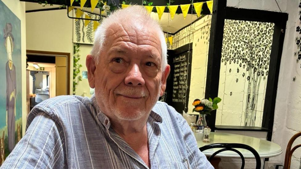 Gatwick Area Conservation Campaign chairman Peter Barclay is wearing a striped shirt and sitting at a table with bunting above him and a picture on a wall next to him.