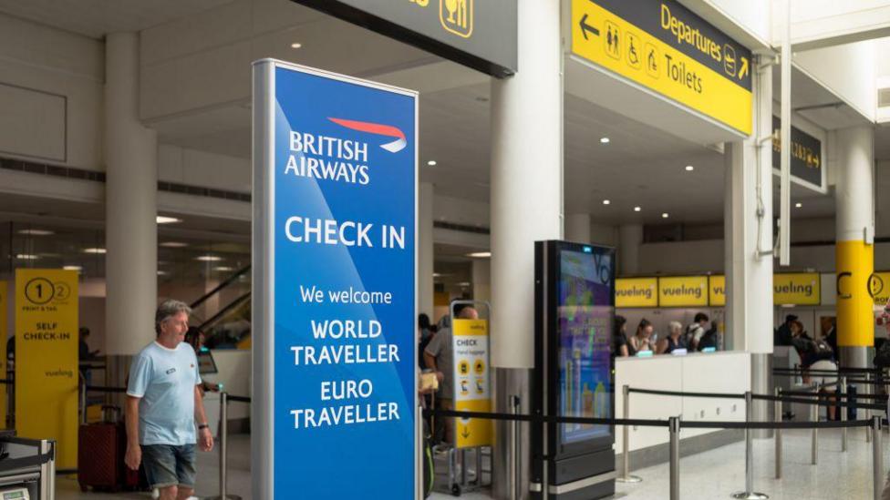 A check in area at London Gatwick has a sign for British Airways. Staff are being a desk and a man in a T-shirt is near a sign that says self check-in. There is a screen near the check-in desks. A yellow sign overhead points to Departures.